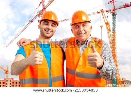Older construction supervisor with younger worker helper at construction site. Managers wearing protective workwear, hard hat, construction cranes on skyline. Construction 