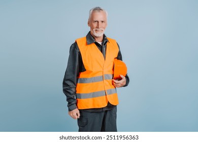 Older construction worker wearing a high visibility vest holding his hard hat is smiling at the camera on a blue background - Powered by Shutterstock