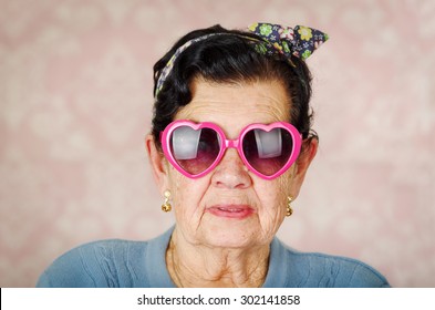 Older Cool Hispanic Woman Wearing Blue Sweater, Flower Pattern Bow On Head And Pink Heartshaped Sunglasses Looking Into Camera.