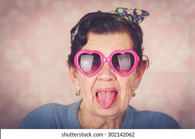 Older Cool Hispanic Woman Wearing Blue Sweater, Flower Pattern Bow On Head And Pink Heartshaped Sunglasses Looking Into Camera Showing Her Tongue.