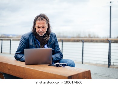 Older but cool man with beard and long grey hair writes on his laptop on the riverbank.