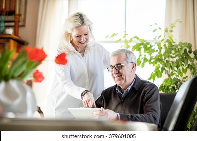 Older Couple Having Fun And Smiling While Working From Home On Notebook With Green Flowers And Window Light Around Them