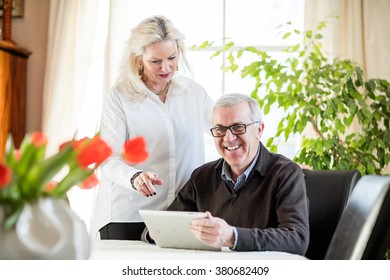 Older Couple Having Fun And Smiling While Working From Home On Notebook With Green Flowers And Window Light Around Them