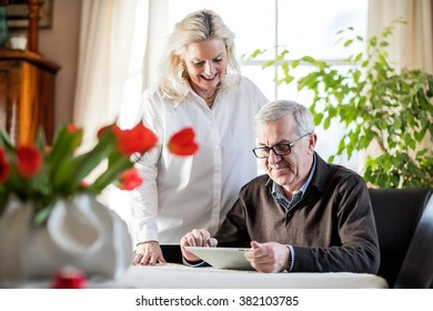 Older Couple Having Fun And Smiling While Working From Home On Notebook With Green Flowers And Window Light Around Them