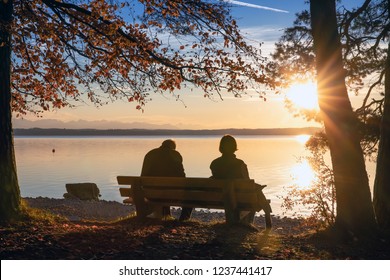 Older Couple Man, Woman Are Sitting On A Park Bench At A Beautiful Sea Lake See And Look Into A Colorful Sundown - In The Background You Can See The Mountains Alps