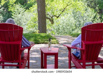 Older couple - man and woman - sitting in red muskoka chairs on backyard patio looking out over yard. Trees in background. - Powered by Shutterstock