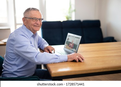 Older Couple Talks To Each Other Via Video Conferencing. Happy Gray-haired Man With Glasses Observes On The Screen His Wife Preparing A Meal In The Kitchen At Home.