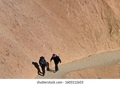 Older Couple Walking Up A Hill Trail Along A Dry Canyon Side.  Both Are Dressed Appropriately For Cool, Sunny Weather