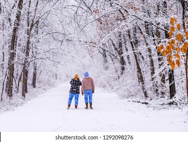 Older Couple Walking On Trail In Beautiful Snowy Park During Snowfall; Woman Is Taking Pictures With Her Smartphone