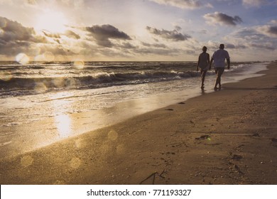 Older couple walks on the beach holding hands at sunset just as a wave retreats from the shore.