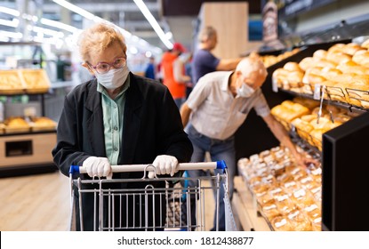 Older European Woman  Wearing Mask And Gloves With Covid Protection Chooses Buns And Bread In Supermarket Bakery