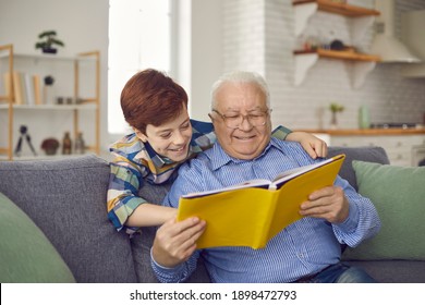 Older Family Members And Children Having Fun Together: Happy Grandfather And Grandson Reading Book Of Good Fairy Tales On Cozy Couch At Home. Senior Man And Little Kid Enjoying Free Time On Weekend