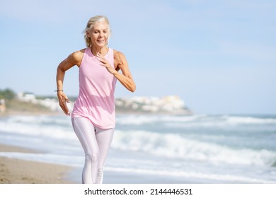 Older Female Doing Sport To Keep Fit. Mature Woman Running Along The Shore Of The Beach. Concept Of Healthy Living In The Elderly. Senior Woman In Fitness Clothing Running Along Beach