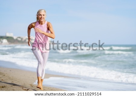 Older female doing sport to keep fit. Mature woman running along the shore of the beach. Concept of healthy living in the elderly. Senior woman in fitness clothing running along beach