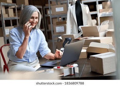 Older female fashion dropshipping business owner using laptop talking on phone in warehouse. Mature woman seller merchant checking online website order confirming ecommerce shipping parcel delivery. - Powered by Shutterstock