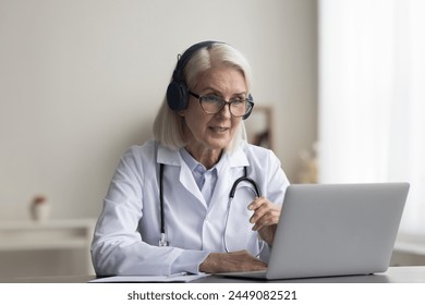 Older female GP sit at desk with modern laptop take part in videocall event, talk to patient, discuss treatment plan, provide counseling or mental health support, tell about preventive care measures - Powered by Shutterstock