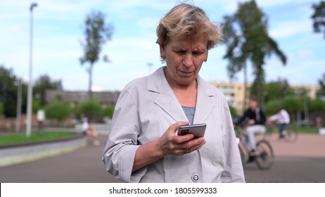 An Older Focused Woman Writes A Message On Her Brand New Smartphone Walking On The Street.