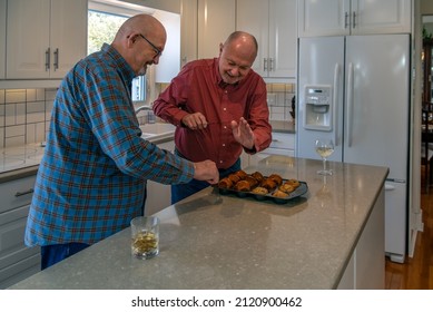 An Older Gay Man Is Playfully Slapping Away His Husband's Hand As His Husband Reaches For A Muffin Fresh Out Of The Oven.