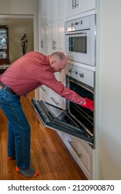 An Older Gay Man Is Reaching Into His Wall Oven To Pull Out A Fresh Baked Batch Of Muffins.