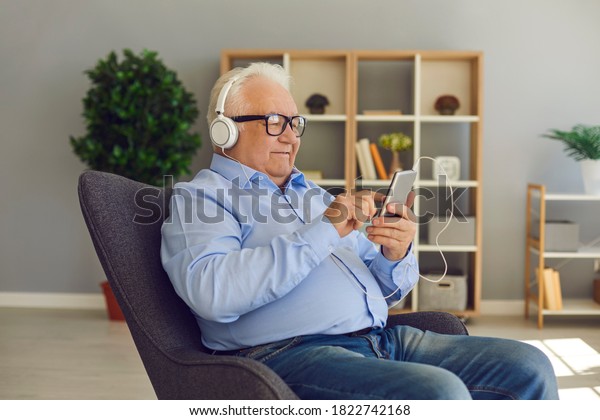 Older generation embraces new technology. Happy\
relaxed senior man with cool headphones and a cell phone listening\
to favorite music or podcast using an easy app sitting in an\
armchair at home