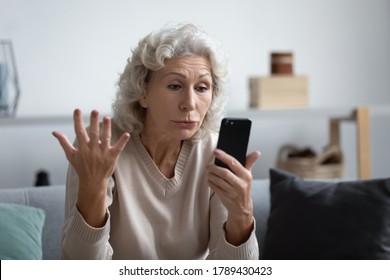 Older Generation And Modern Technology Problems Of Usage, Not Understand Need Help Concept. Old Grey Haired Woman Sit On Sofa Holds Smart Phone Looks At Device Screen Gesturing Feels Annoyed And Angry