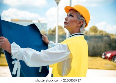 Older Gray Hair Woman Architect With Blueprint Controlling Construction Site. Elderly Female Civil Engineer With Yellow Hardhat And Reflective Vest Work As Supervisor