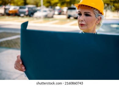 Older Gray Hair Woman Architect With Blueprint Controlling Construction Site. Elderly Female Civil Engineer With Yellow Hardhat And Reflective Vest Work As Supervisor. Copy Space