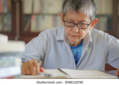 Older Gray Hair Woman Thoughtfully Handwrote Testament. Senior Asian Woman Sitting In Home Book Library Taking Notes On A Notebook, Writing Diary Or Biography. Elderly People Carefully Prepare A Will.