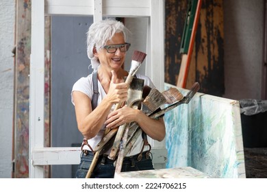 older gray haired mature happy artist woman with glasses and big paintbrushes gives thumbs up, copy space - Shutterstock ID 2224706215