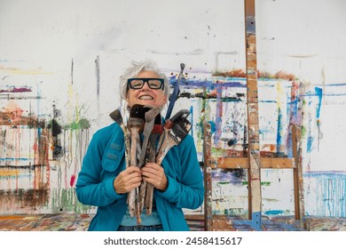 older gray haired mature happy smiling artist woman proud artist, in her fifties with grey hair and glasses and big paintbrushes, in her studio, copy space - Powered by Shutterstock