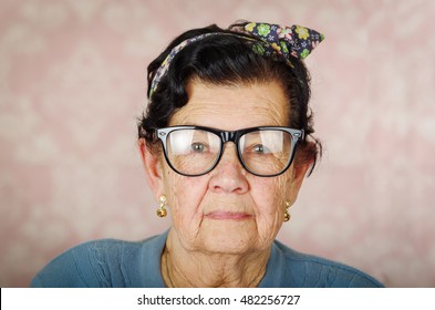 Older Hispanic Cute Woman With Flower Pattern Bow On Her Head Wearing Blue Sweater And Black Large Framed Glasses Looking Into Camera