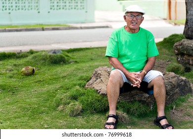 A Older Hispanic Senior Citizen Man Sits Outdoors In A Tropical Setting.