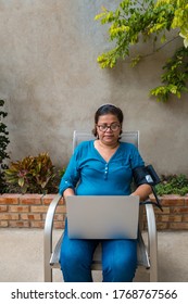 An Older Hispanic Woman Checking Her Blood Pressure And Pulse At Home. The Retired Woman Sits In Front Of A Laptop Monitor And Measures Her Vital Signs