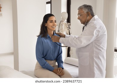 Older Indian doctor man checking heartbeat rate of young patient, applying stethoscope to chest, speaking, asking about symptom. Positive Latin woman visiting cardiologist for checkup - Powered by Shutterstock