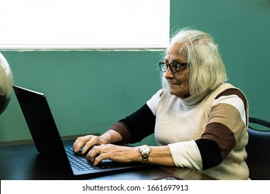 Older Lady With Glasses Sitting Typing On A Black Laptop, With A Green Wall In The Background