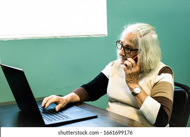 Older Lady With Glasses Sitting Typing On A Black Laptop I Talking With A Cellphone, With A Green Wall In The Background