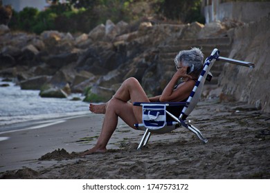 Older Lady Sitting In A Chair On The Beach Listening To The Phone