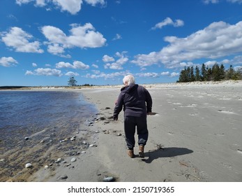 An Older Lady Walking Along The Water On A Sandy Beach.