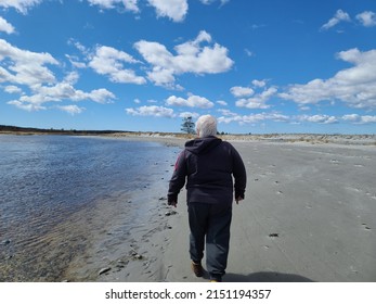 An Older Lady Walking Along The Water On A Sandy Beach.