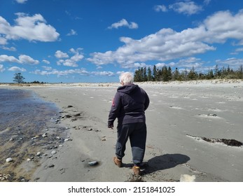 An Older Lady Walking Along The Water On A Sandy Beach.