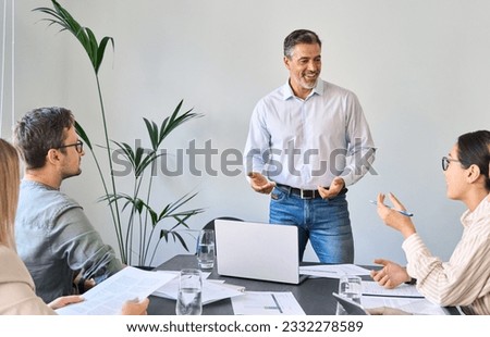 Older Latin company manager leading team meeting in office. Happy diverse professional people group working together in boardroom. Mature business man boss talking to colleagues having discussion.