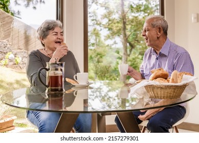 An older Latino man and woman sitting in the dining room of their home chatting and drinking coffee.