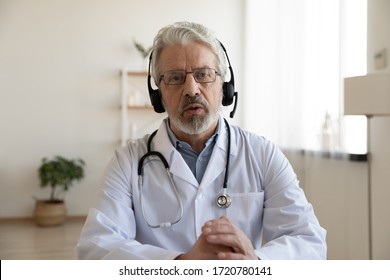 Older male doctor therapist wearing headset videoconferencing talking to web camera consulting virtual patient online by video conference call chat. Telemedicine, telehealth concept. Webcam view.