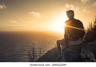 Older male hiker sitting relaxing on the edge of mountain cliff facing spectacular views of the ocean sunrise. Senior people adventure travel, independence concept. 