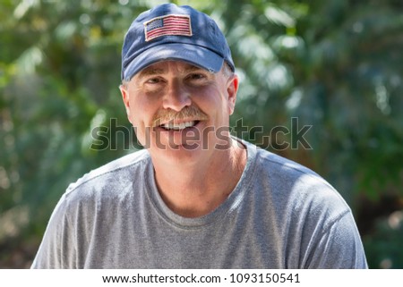 Older male smiles warmly outside wearing USA flag baseball hat