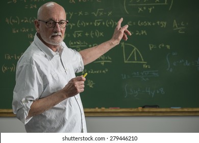 Older Male Teacher Standing In Front Of A Chalkboard In A Classroom