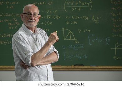 Older Male Teacher Standing In Front Of A Chalkboard In A Classroom