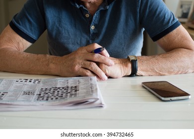 Older Man In Blue Shirt Leaning On Table Holding Pen With Crossword And Phone In Front Of Him (cropped And Selective Focus)