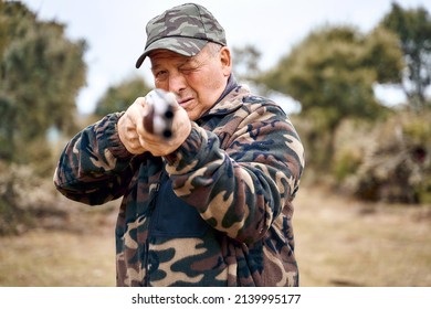 Older Man In Camouflage Clothing And Cap Squinting And Pointing Shotgun At Camera While Hunting In A Field On An Autumn Day. 