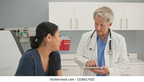 Older man doctor listening to medical history from Japanese woman in exam room while holding tablet computer. Senior Caucasian male medical professional in OBGYN office with patient